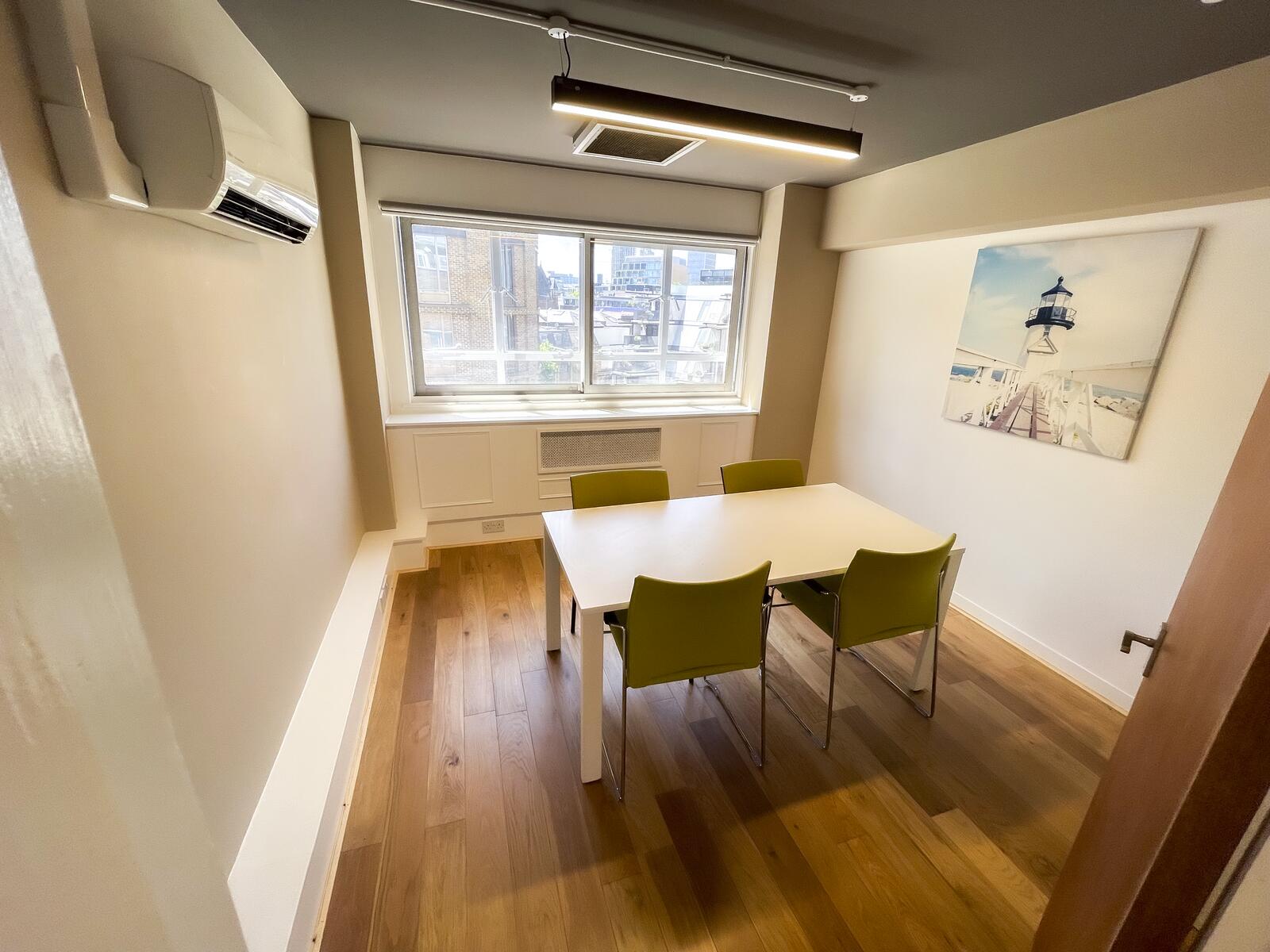 Compact fourth-floor meeting room with a white table and four green chairs, wooden flooring, large window for natural light, wall-mounted air conditioning, and modern overhead lighting—ideal for private discussions or small team meetings.