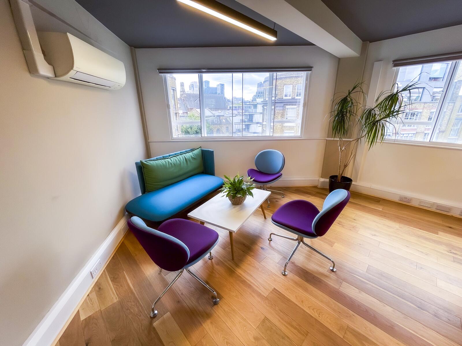 Bright breakout seating area with a sofa, purple chairs, and a white coffee table, set on wooden floors beside large windows with city views, indoor plants, and modern ceiling lighting.