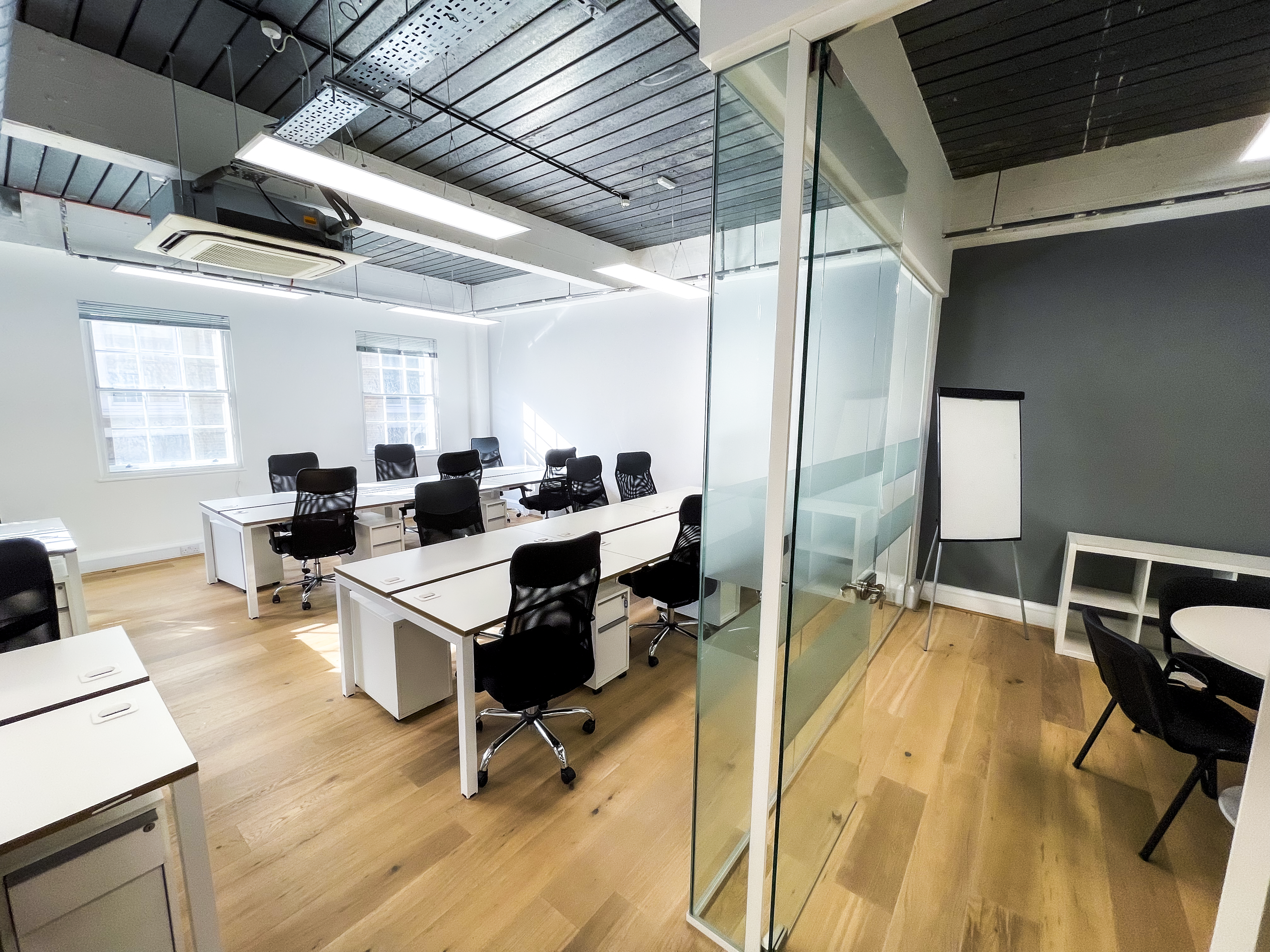 Open-plan office with rows of white desks and black ergonomic chairs, wooden floors, and large windows for natural light, alongside a glass-walled meeting room with a flip chart and small table.