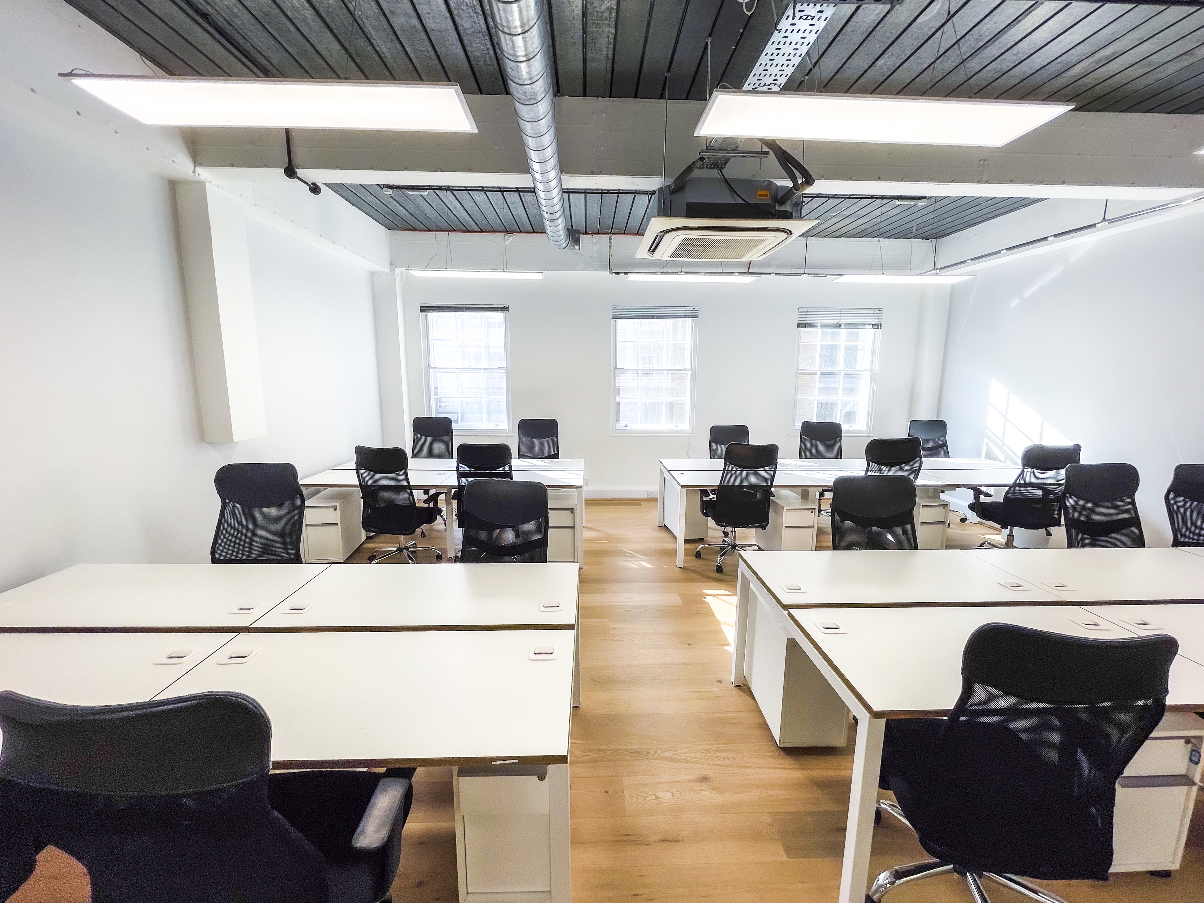 Open-plan office with white desks and black ergonomic chairs arranged in rows, wooden floors, three large windows providing natural light, and an exposed industrial ceiling with LED panels and air conditioning.