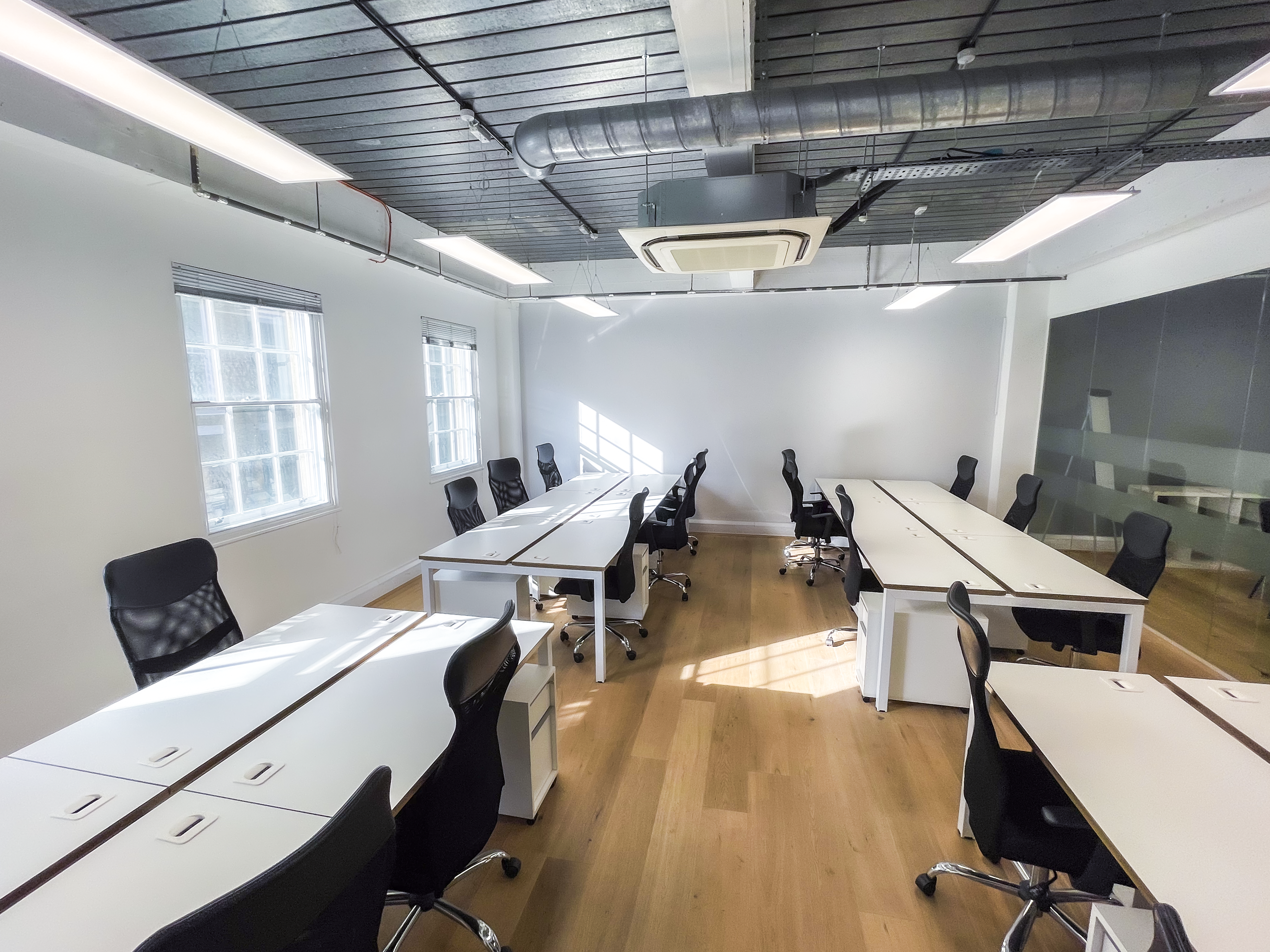 Bright open-plan office with rows of white desks and black ergonomic chairs, wooden floors, large windows letting in natural light, and an exposed industrial ceiling with modern lighting and air conditioning.