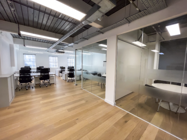 Open-plan office with wooden floors, rows of white desks and black chairs, and two glass-walled meeting rooms on the right, allowing privacy while keeping the space bright and connected.