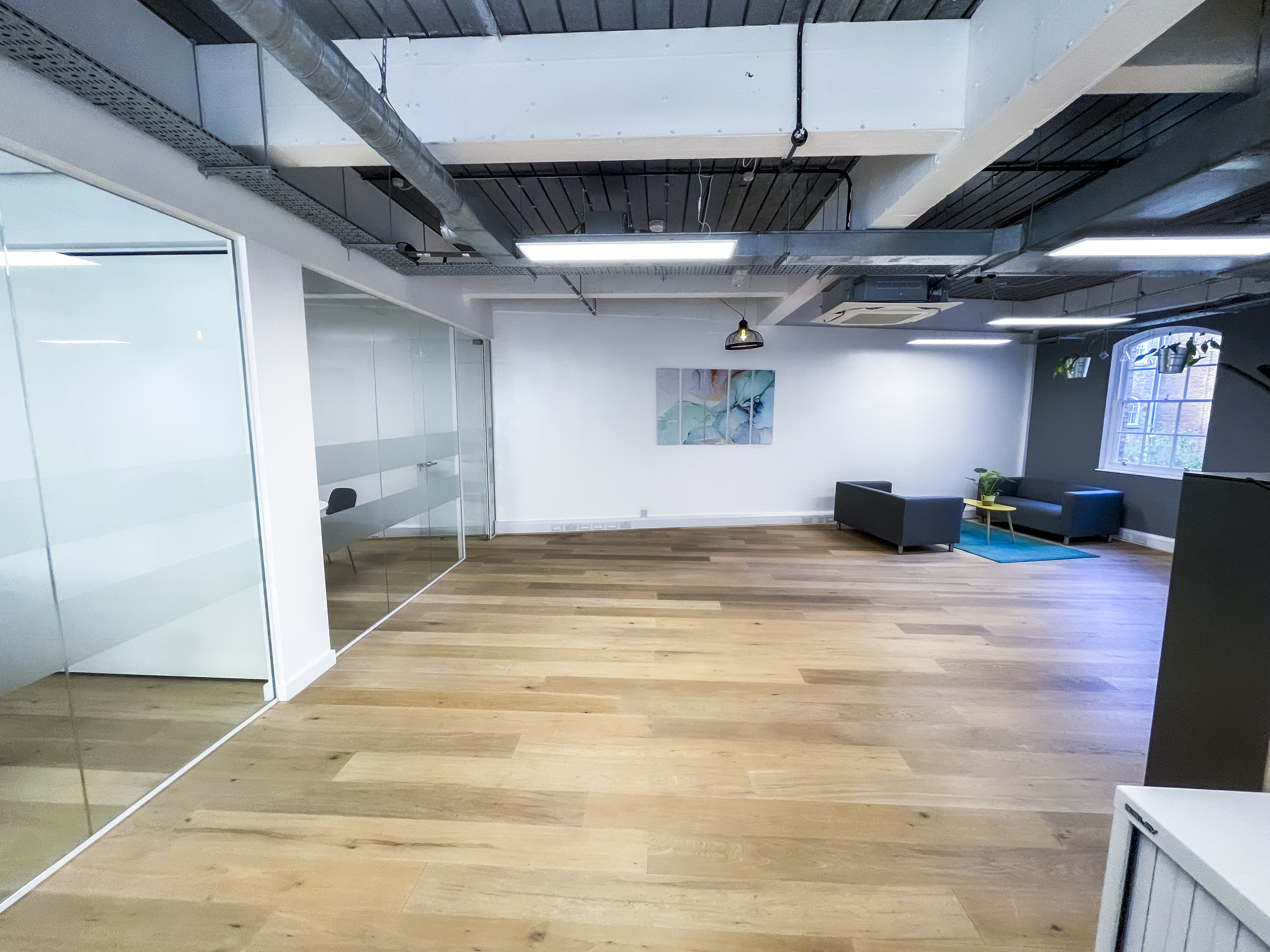 Spacious second-floor office area with wooden floors, glass-walled meeting rooms on the left, exposed industrial ceiling, and a small lounge corner with sofas, plants, and artwork on the far wall.