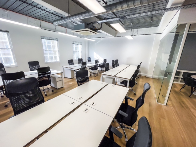 Open desk area with rows of white workstations and black ergonomic chairs, wooden floors, large windows for natural light, and a partial view of a glass-walled meeting room on the right.