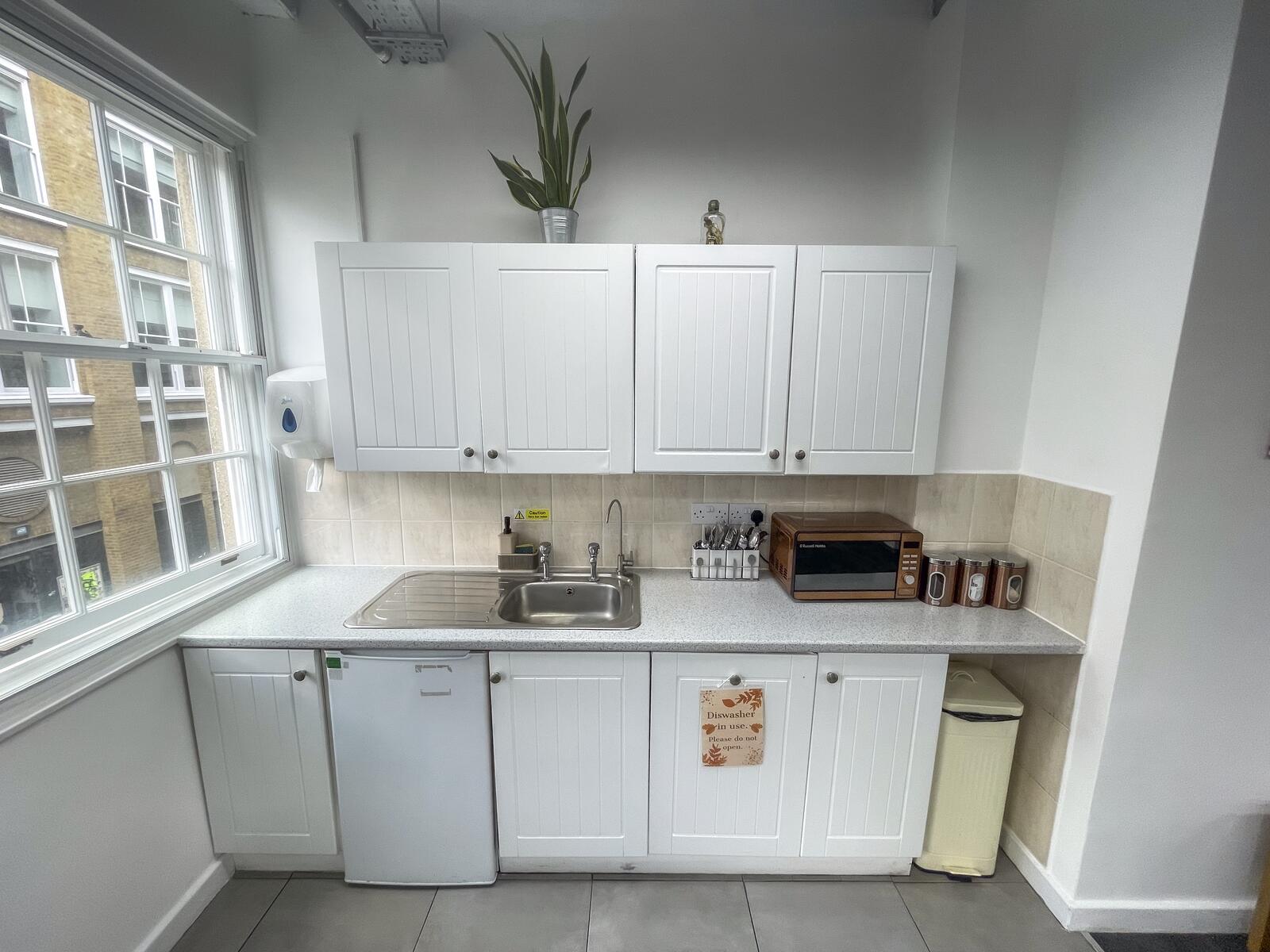 Second-floor private office kitchenette with white cabinets, sink, compact fridge, microwave, storage jars, and a large window bringing in natural light.