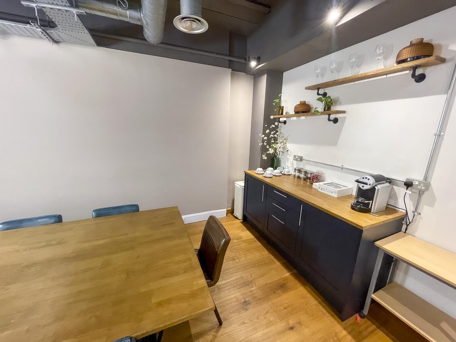 The Orbit meeting room with a wooden table and chairs beside a kitchenette featuring black cabinets, a coffee machine, open shelves with glassware and plants, and warm wooden flooring under an industrial ceiling.