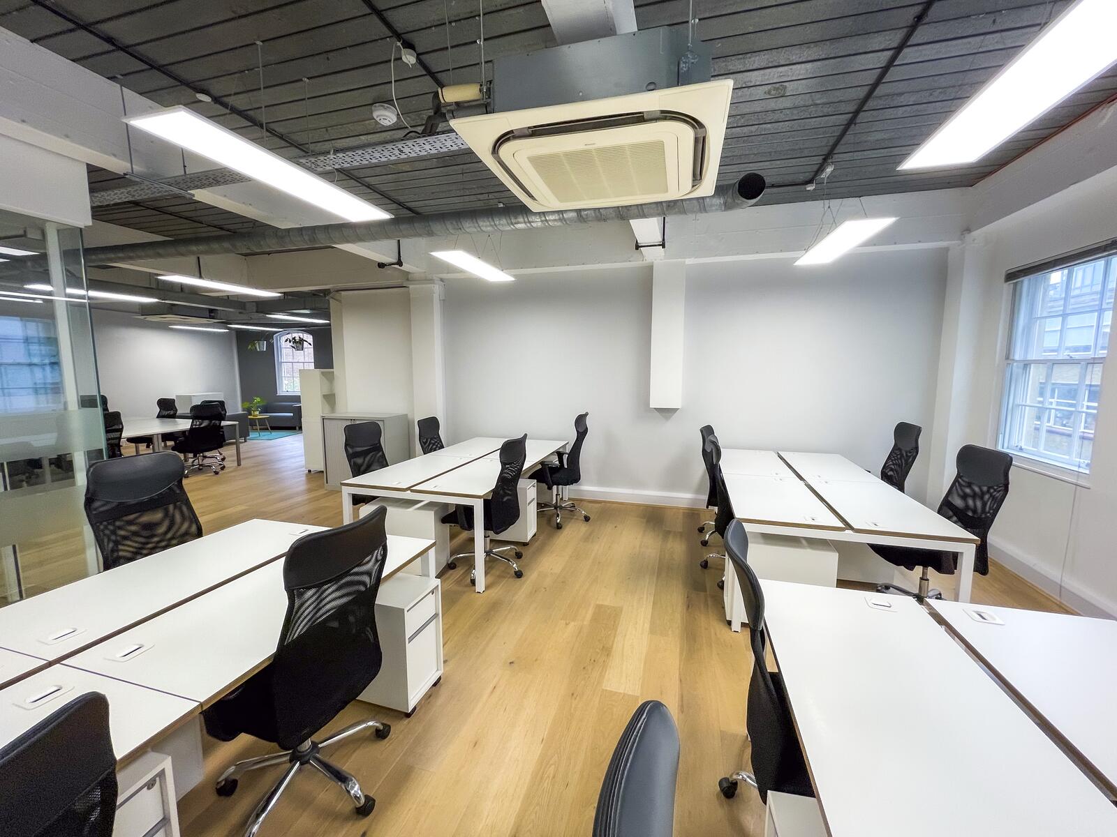 Open-plan second-floor office with white workstations, black ergonomic chairs, wooden floors, large windows for natural light, and exposed ceiling with modern lighting and air conditioning.