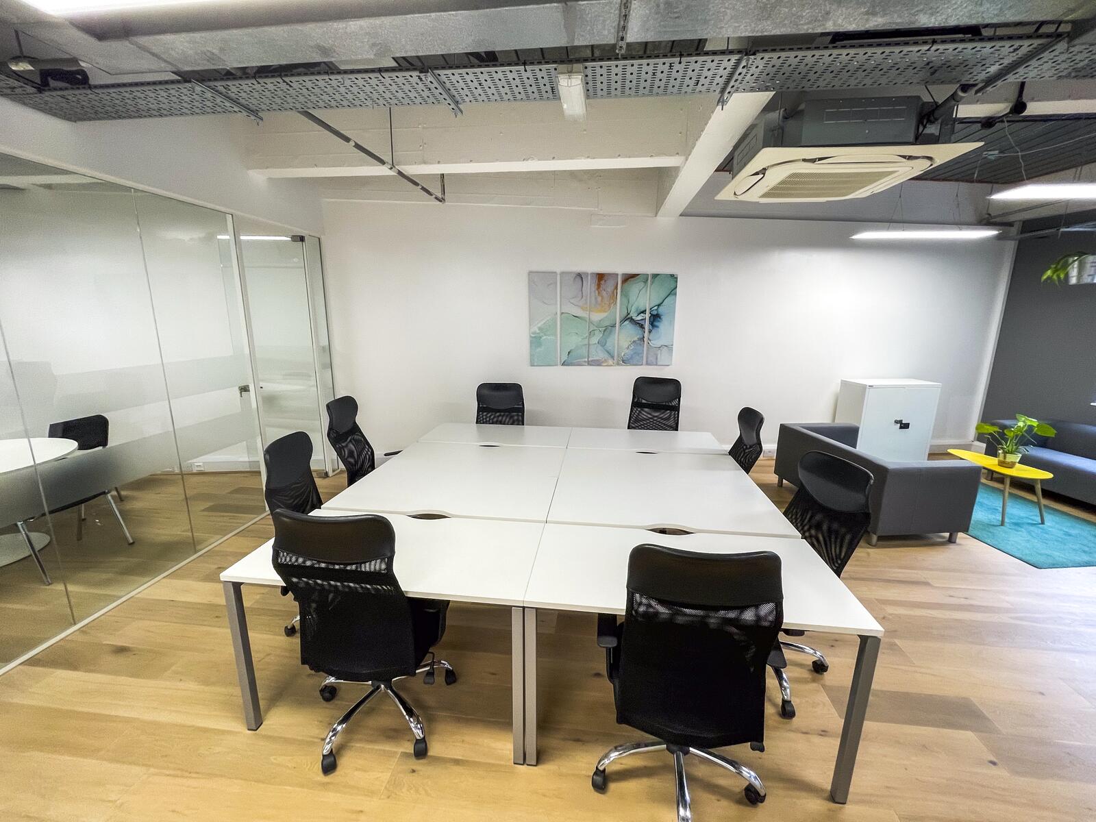 Second-floor office with an open-plan desk area, white workstations and black ergonomic chairs, two glass meeting rooms, wooden floors, and a small lounge space with sofas and plants.